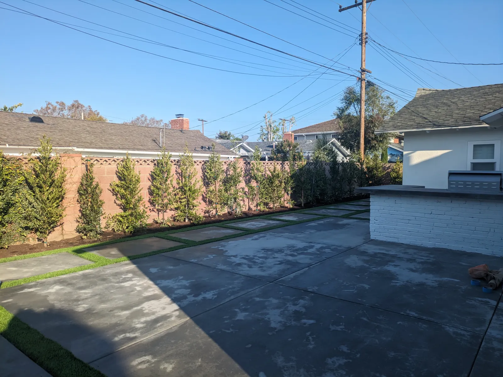 Fresh concrete slab patio featuring green turf strip inserts alongside a line of hedge trees.