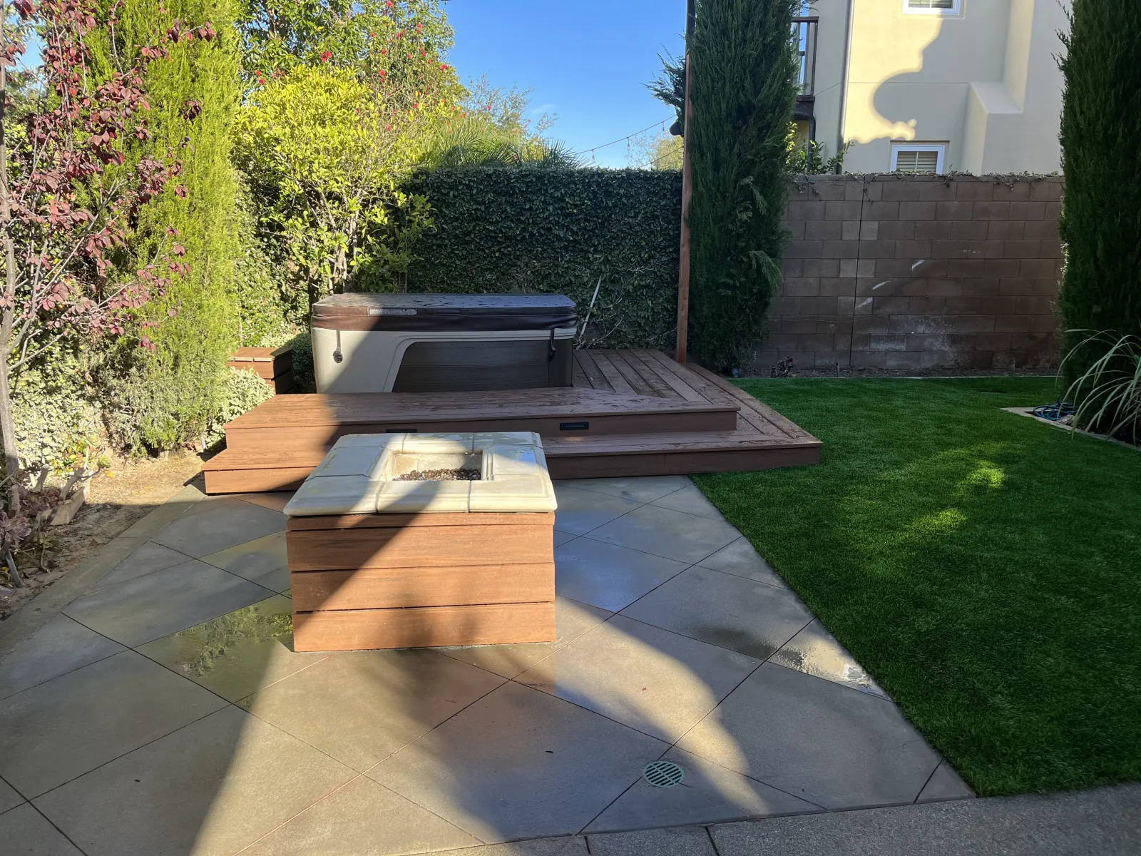 Backyard patio showing a square wooden fire pit in the foreground, flagstone patio flooring, and a raised wooden deck with a hot tub in the background.