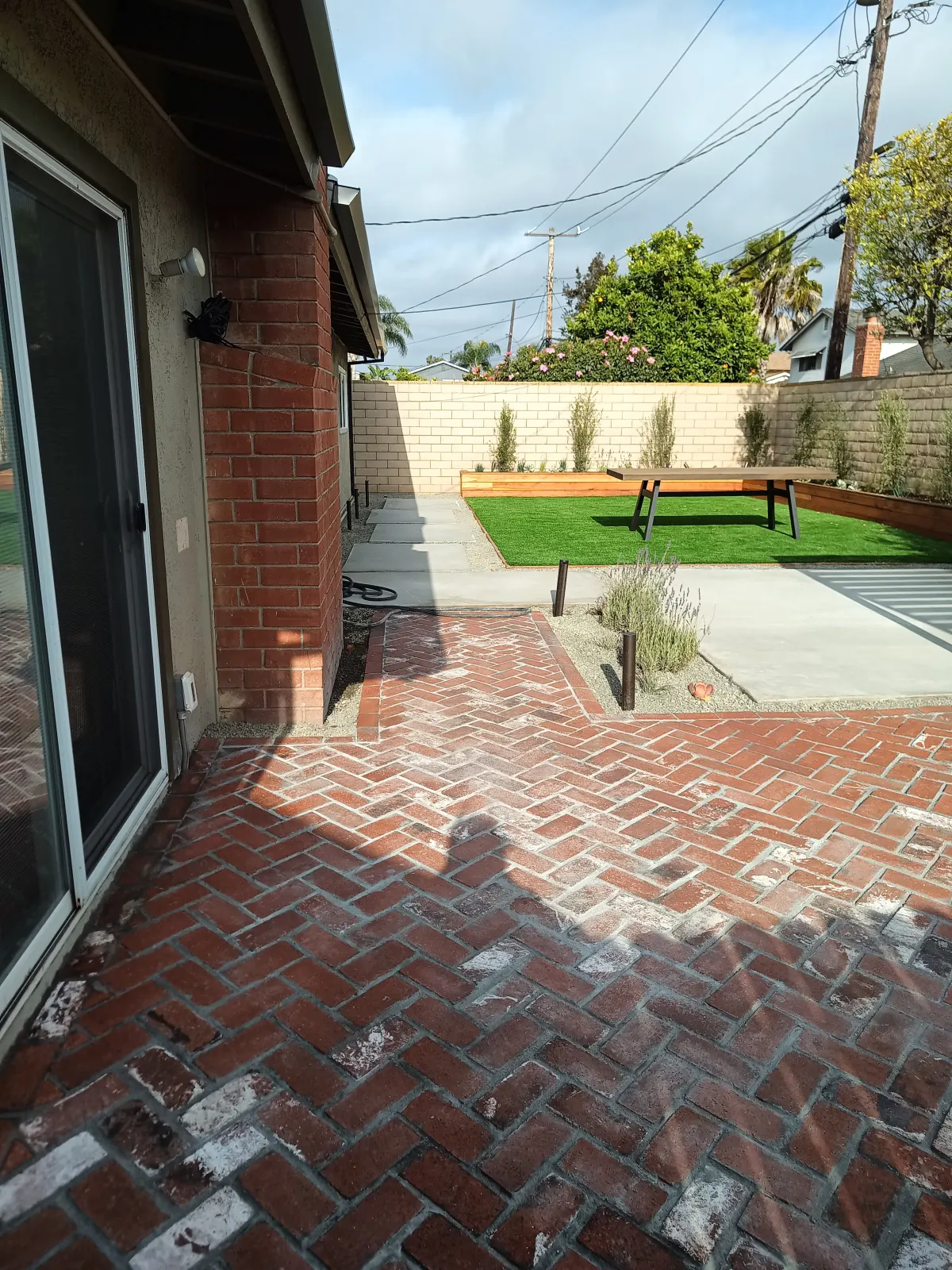 View from a patio showing red brick paving in a herringbone pattern, leading out towards a sunny side yard with artificial turf.